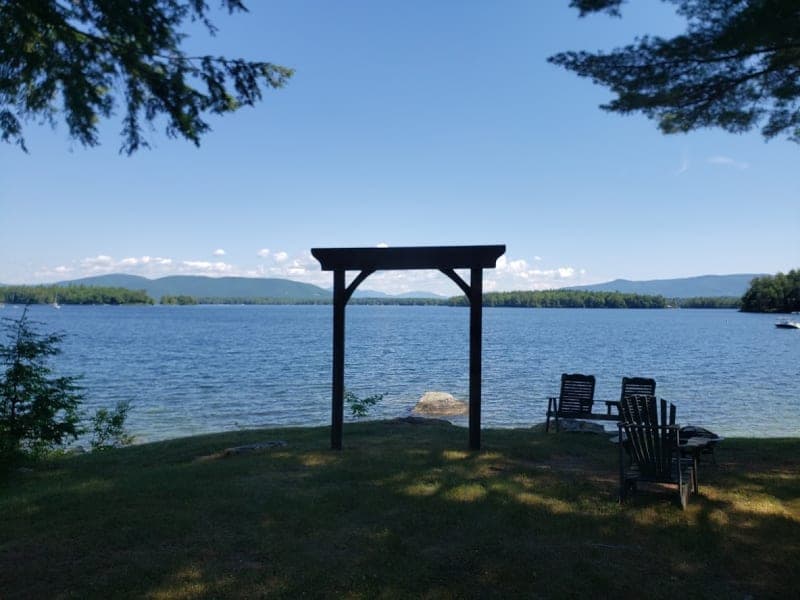 A serene lake view featuring a wooden arch and two wooden chairs on the grassy shore.