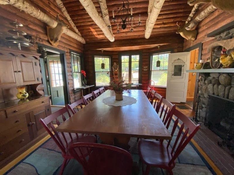 A rustic dining room with a large wooden table surrounded by red chairs, wooden beams, and stone fireplace.