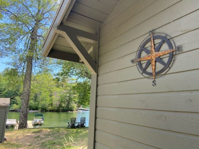 A compass mounted on the side of a cabin overlooks a tranquil lake with wooden chairs by the shore.