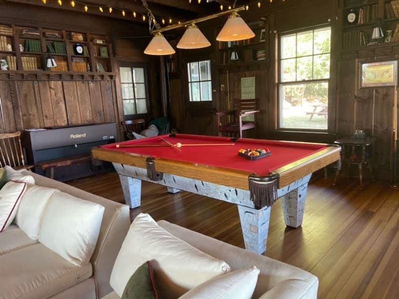 A cozy game room features a red felt pool table, comfortable seating, and wooden shelves filled with books.